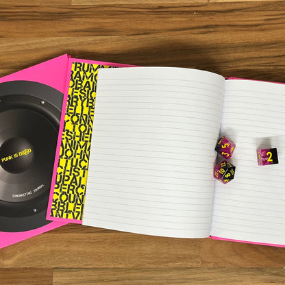 Open notebook with dice and a pink and black object on a wooden surface